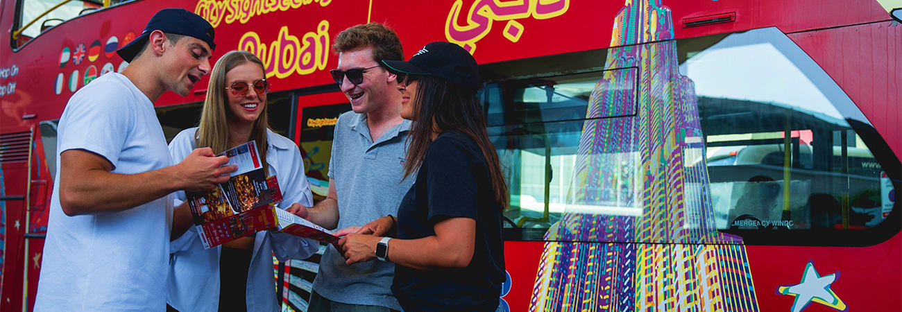 Group of tourists receiving tour information from guide next to red double-decker sightseeing bus with colorful city skyline 